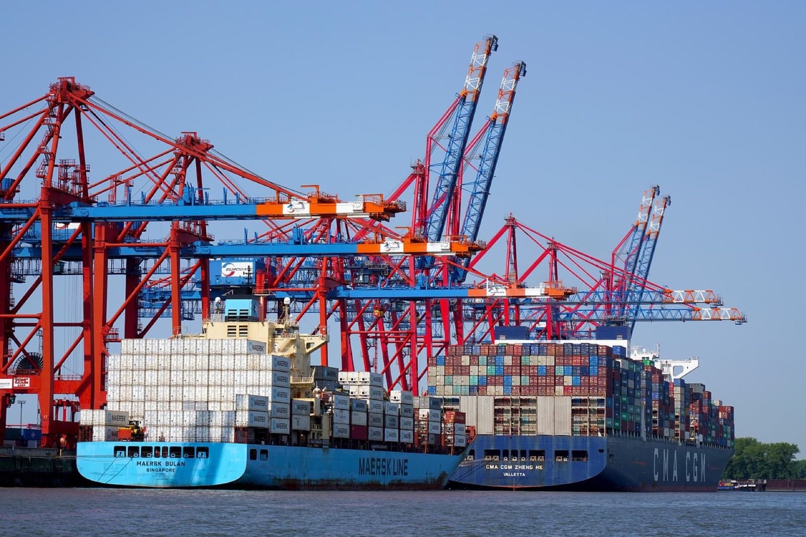 Container ships alongside a quay under red and blue gantry cranes, stacked containers being loaded.