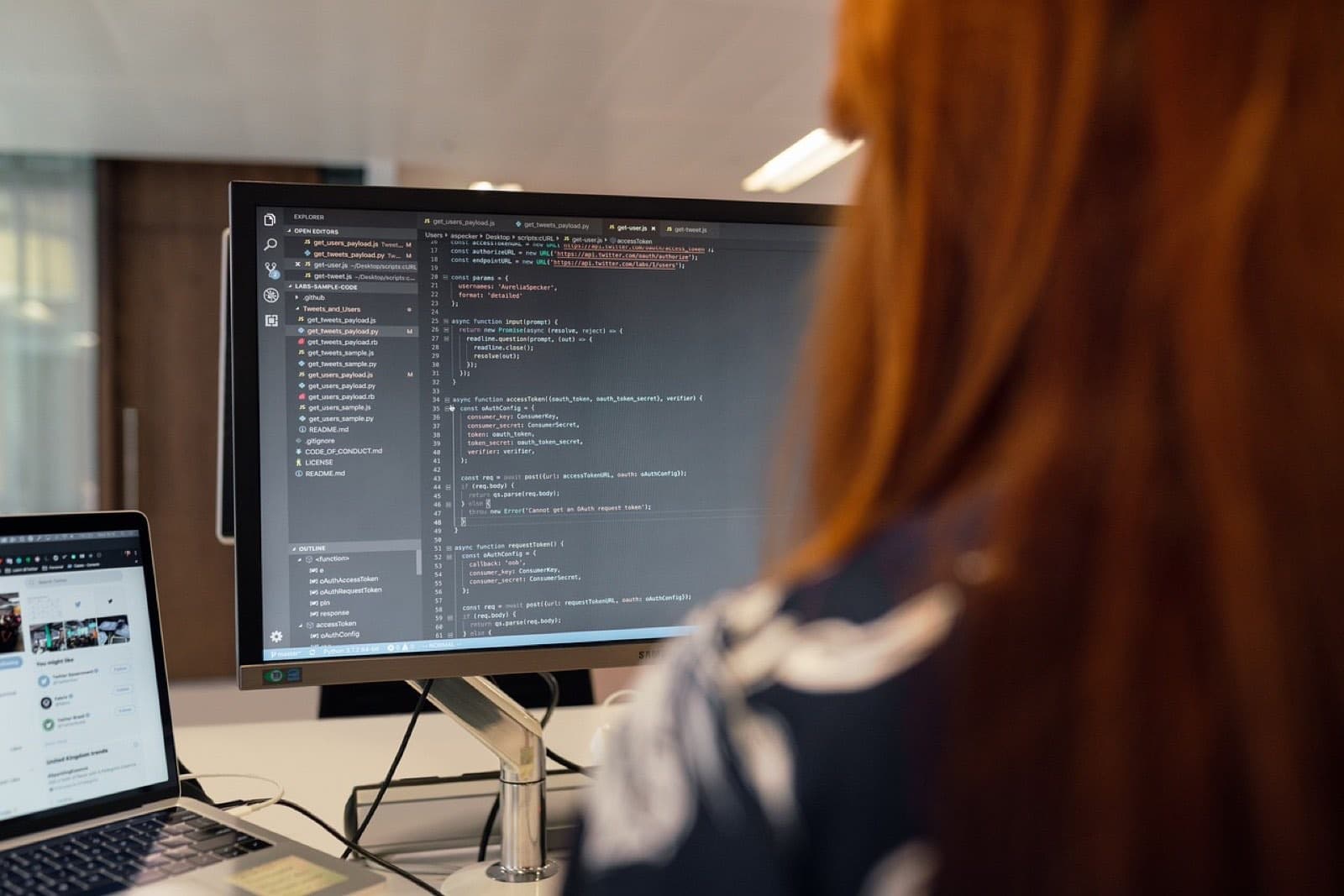 An engineer at a desk reading code on a large monitor, a laptop open beside it.